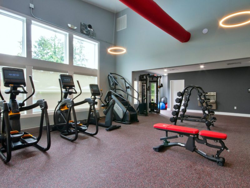 A picture showing the workout athletic club showing stair steppers, stationary bike, front of windows, a bench, and free weights and workout station with a drinking fountain on the wall.